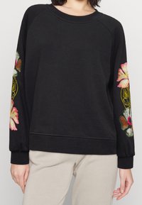 Sudadera negra con bordado floral en rosa y blanco en las mangas, cuello redondo y dobladillo acanalado. Tejido suave con un corte holgado.