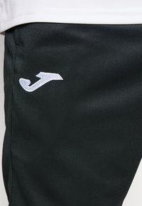 Sort atletikbukser med en glat tekstur, der har et hvidt broderet logo på siden. Simpelt design med en diskret silhuet.