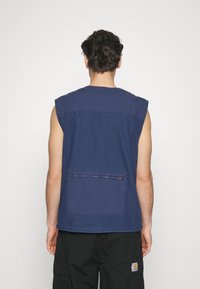 Bezrękawnik dżinsowy w ciemnoniebieskim kolorze, z kwadratową kieszenią z tyłu, z zamkiem błyskawicznym i przeszyciami. Gładka faktura.