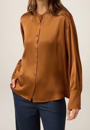 Mujer con una blusa de satén camel marrón, suelta, de manga larga y con botones, y pantalones azul oscuro, de pie con una mano en el bolsillo.