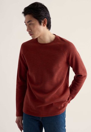 Homme aux cheveux noirs portant un pull à manches longues couleur rouille et un jean bleu, regardant vers sa gauche avec une main dans sa poche.