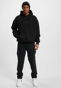 Schwarzer übergroßer Kapuzenpullover mit einer Vordertasche, kombiniert mit schwarzen Jeans und weißen Sneakern. Texturiertes Gewebe und ein einfaches, klares Design.
