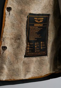 Capuchonjack met een zachte, beige fleece voering, zwarte buitenlaag en een productlabel met specificaties en verzorgingsinstructies.