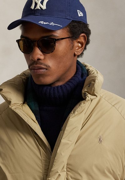 Bēša pūķveida jaka ar augstu apkakli, ar izšūtu logotipu uz krūtīm, nēsāta pāri tumši zilam augstajam apkaklam, papildināta ar apaļām bruņurupuču krāsas saulesbrillēm un zilu cepuri.