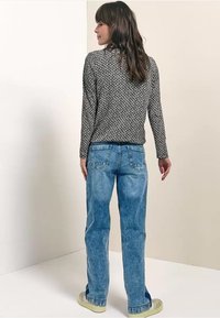 Donna con capelli castani lunghi che indossa una maglia a maniche lunghe con motivo bianco e nero, jeans blu a vita alta e sneakers beige, di spalle.