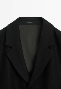 Blazer en laine noire avec un col à revers aiguisé, un tissu texturé et une doublure sombre. Étiquette visible indiquant "FABRIQUÉ EN ÉGYPTE".