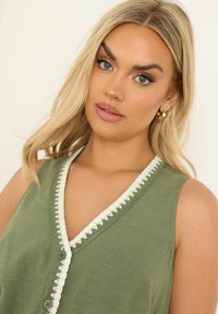 Oliivivihreä hihaton toppi, jossa on v-aukko. Topissa on valkoista virkattua reunusta ja kaksi nappia edessä. Pehmeä kangas ja rento istuvuus.