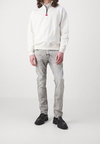 Biała bluza na zamek z wysokim kołnierzem, w połączeniu z jasnoszarymi, przetartymi dżinsami i czarnymi mokasynami. Prosty, codzienny styl.