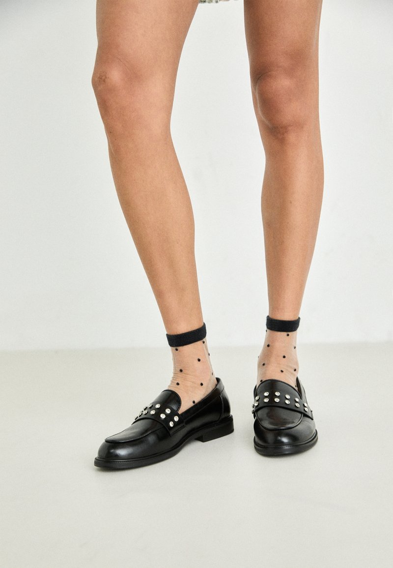 Sorte skinnloafers med sølvnagler, kombinert med gjennomsiktige sokker med prikker. Loaferne har en rund tå og flat såle.