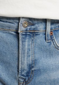 Jeans de denim azul claro con cierre de botón, cremallera y costuras contrastantes a lo largo de las costuras. Desvanecimiento sutil y textura evidente.