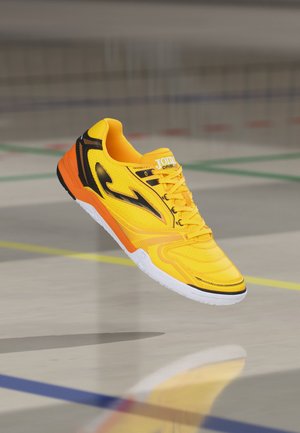 Sárga és fekete színű, fehér talpú beltéri sportcipő, amely egy edzőtermi padló felett lebeg, amelyet különböző sportágak színes vonalai jeleznek.