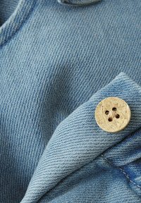 Jasnoniebieski materiał jeansowy o widocznej fakturze, z drewnianym guzikiem z czterema dziurkami na złożonym fragmencie materiału.