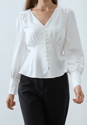 Mujer con una blusa blanca a medida con botones de tela y mangas largas abullonadas, combinada con pantalones negros de cintura alta.