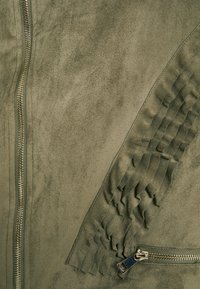 Veste en faux daim vert olive avec une fermeture éclair diagonale, détail plissé texturé sur les manches, et quincaillerie de fermeture éclair métallique.