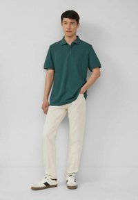 Polohemd aus dunkelgrüner Baumwolle, kurzärmelig, lockere Passform. Kombiniert mit weißen Hosen und Sneakers in Schwarz und Beige.