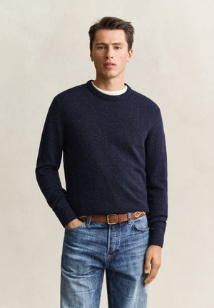 Young man wearing a dark blue speckled sweater, brown belt, and blue jeans standing with one hand in pocket against a plain light background.