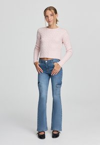 Pullover cropped in maglia cable rosa chiaro abbinato a jeans flare blu a vita alta con tasche cargo e ballerine nere.