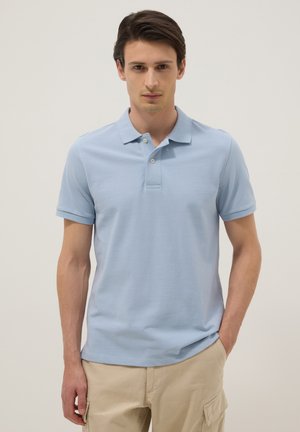 Junger Mann, der ein hellblaues Poloshirt und beige Cargo-Shorts trägt, steht mit einer Hand in der Tasche vor einem einfarbigen Hintergrund.