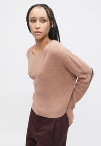 Světle růžový žebrovaný pletený svetr s dlouhými rukávy a širokým kulatým výstřihem, kombinovaný s tmavě vínovými kalhotami. Měkká textura, boční rozparky na lemu.