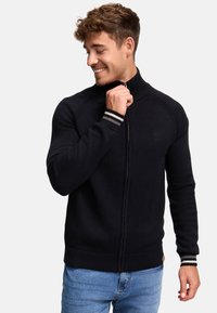 Pull à zip noir avec col et poignets côtelés. Texture tricot, design ajusté et un petit logo sur la poitrine. Associé à un jean bleu.