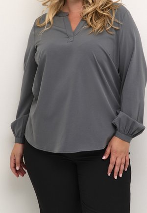 Kvinde iført en grå bluse med lange ærmer med rynkede manchetter og sorte bukser, stående mod en ensfarvet hvid baggrund.