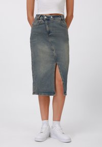 Jupon en jean bleu clair avec une fente avant, taille haute et deux boutons en haut. Porté avec des baskets blanches et des chaussettes visibles.