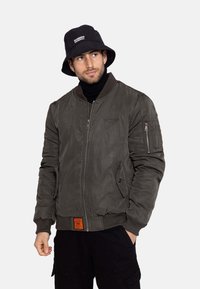 Olivenfarbene Bomberjacke aus glattem Material, mit einem Reißverschluss vorne, Seitentaschen, gerippten Bündchen und einem orangefarbenen Etikett am Saum.