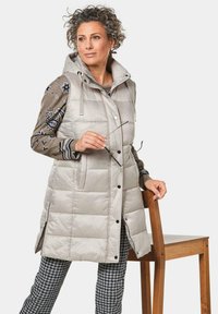 Hellegraue gepolsterte Weste mit Kapuze, die Druckknöpfe, Seitentaschen und gestreifte Besätze hat. Über einem gemusterten Pullover und karierten Hosen getragen.