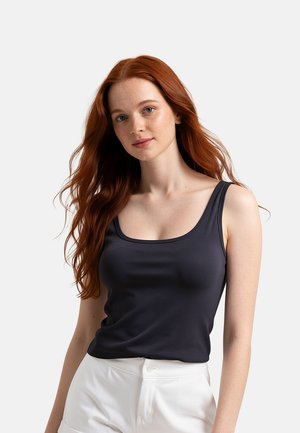 Giovane donna con lunghi capelli rossi che indossa un top nero senza maniche e pantaloni bianchi, in piedi davanti a uno sfondo semplice.