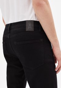 Jean en denim noir avec une finition texturée, une coupe ajustée et une étiquette rectangulaire au dos avec un logo. Comprend deux poches arrière.