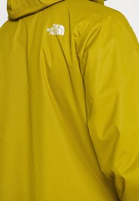 Chaqueta impermeable amarilla con un acabado texturizado, que presenta una capucha y un logo blanco en el hombro. Enfocada en la practicidad y durabilidad.