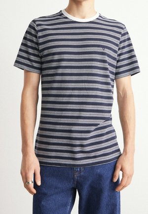 Personne portant un t-shirt à manches courtes rayé horizontalement bleu marine et blanc avec col blanc et jean bleu, debout les mains détendues.