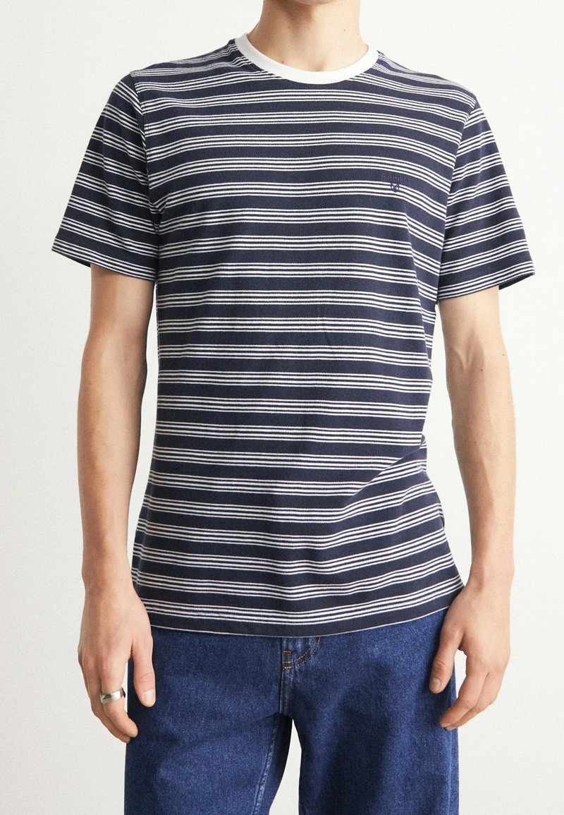 Personne portant un t-shirt à manches courtes rayé horizontalement bleu marine et blanc avec col blanc et jean bleu, debout les mains détendues.