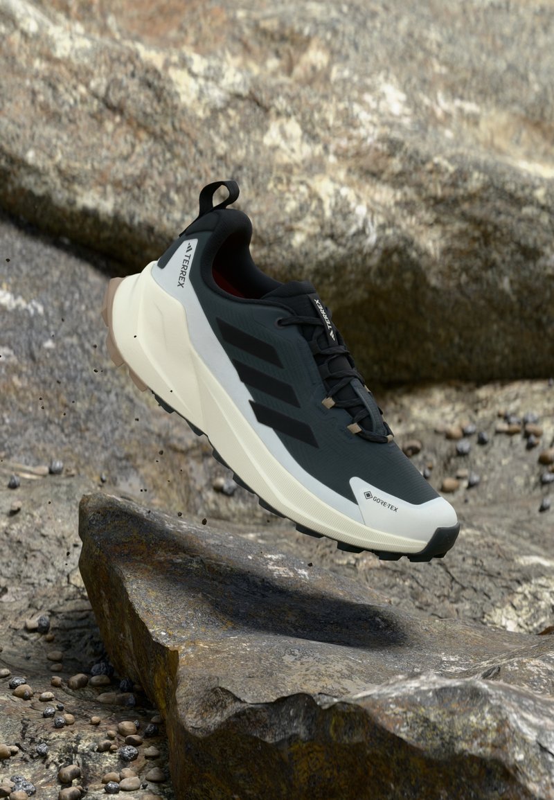 Μαύρα και γκρίζα παπούτσια εξωτερικού χώρου με υφή στο επάνω μέρος, που διαθέτουν τρεις μαύρες ρίγες, λευκή ενδιάμεση σόλα και σήμανση Gore-Tex.