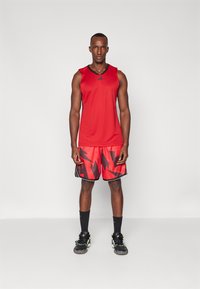 Camiseta sin mangas atlética roja con detalles negros, y pantalones cortos rojos con audaces patrones geométricos negros. El modelo lleva zapatillas y calcetines negros.
