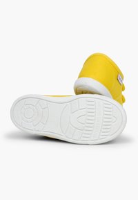 Kollane kanvas sneaker valge kummitald ja tekstuursete aktsentidega, varustatud Velcro rihmaga ja küljepeal oleva sildiga.