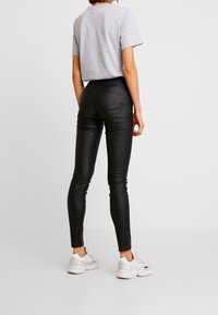 Leggings de cuero sintético negros con una cintura alta, que presentan una textura suave y dos bolsillos traseros, combinados con una camiseta gris de cuello redondo y zapatillas blancas.