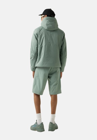 Person wearing sage green hooded jacket, matching shorts, white socks, green sneakers, and black cap, standing facing away on plain background.