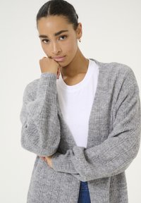Grauer gestrickter Cardigan mit geripptem Textur und offenem Frontdesign. Wird über einem weißen T-Shirt getragen, das lange Ärmel und eine lockere Passform hat.