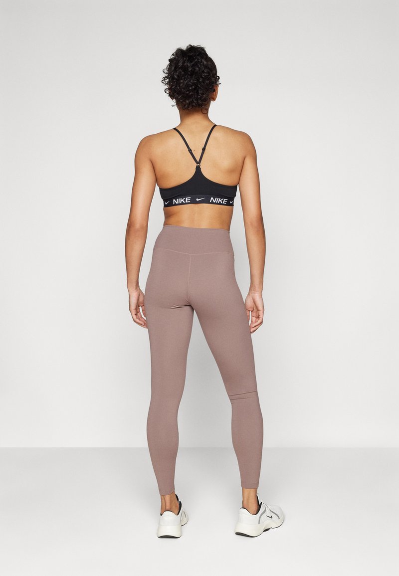 Zwarte sportbra met een logoband, gecombineerd met hooggewrongen, aansluitende taupe leggings. Schoeisel bestaat uit witte sneakers met zwarte accenten.