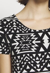 Haut à manches courtes à motif géométrique noir et blanc avec des triangles et des formes en zigzag porté par une personne aux cheveux courts et bruns.