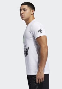Camiseta blanca de manga corta con texto negro y gráficos verdes. Presenta un logotipo en la manga izquierda y un diseño ajustado. Se llevan pantalones cortos negros.