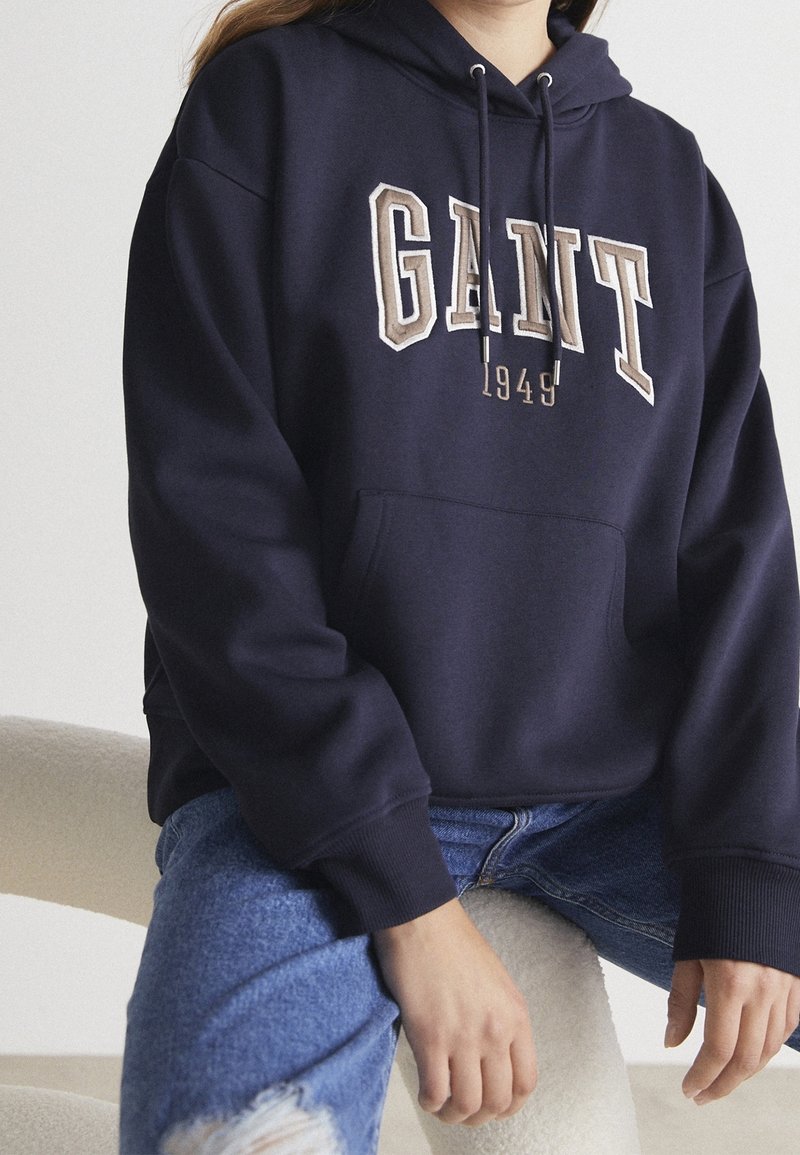 Personne portant un sweat à capuche GANT bleu marine avec des lettres beiges et un jean bleu déchiré, assise sur une chaise claire texturée.