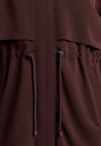 Bordó dzseki, laza szabással, húzózsinóros derékkal és sima textúrával. Részletei között szerepelnek elasztikus díszítések és cipzáras zsebek.