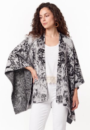 Frau mit lockigem Haar, die einen schwarz-weiß gemusterten offenen Poncho über einem weißen Top und weißen Hosen trägt, posiert mit der Hand in der Hüfte.