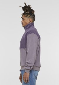 Chaqueta de forro polar en tonos púrpuras, con un alto cuello, cremallera frontal de media longitud y bolsillos laterales. Textura suave con un diseño de paneles en contraste.