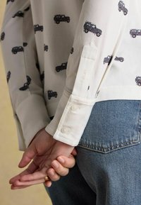 Biela košeľa s čiernym potlačeným autom, s gombíkovými manžetami a viditeľnou značkovou etiketou. V kombinácii s svetlomodrými džínsami.