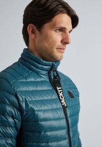 Veste matelassée teal avec col montant, dotée d'une fermeture éclair noire, d'un logo sur la sangle et d'une texture lisse. Design minimaliste avec une coupe ajustée.