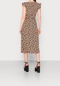 Femme portant une robe midi noire à motifs floraux avec des manches volantes et des sandales à talons noirs, se tenant dos à un fond uni.
