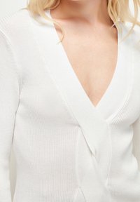 Cardigan blanc à côtes avec un décolleté en V profond. Présente des manches longues et une silhouette ajustée, mettant en valeur un tricot lisse et texturé.